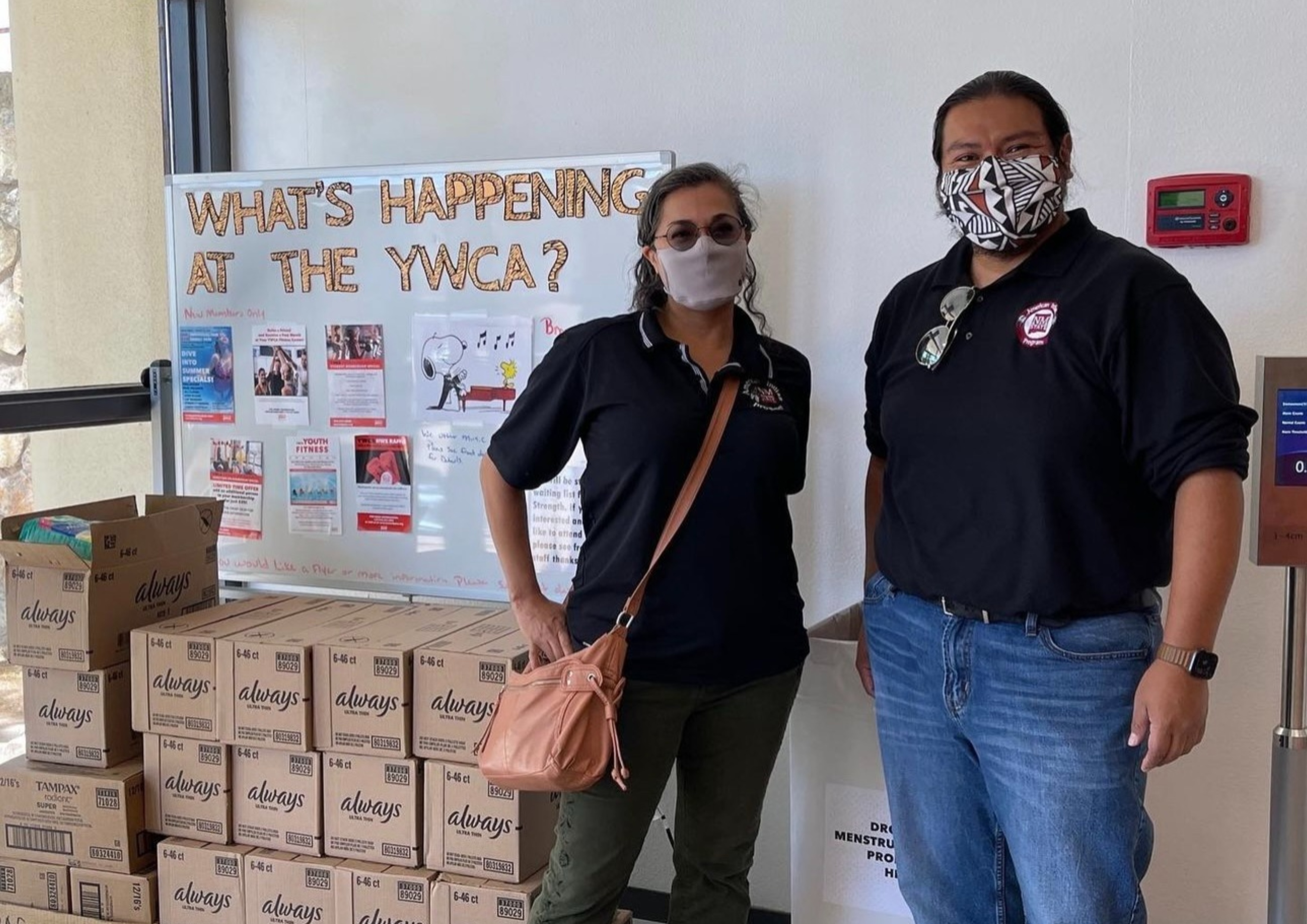 AIP Staff Donating to YWCA. Two masked individuals standing in front of a whiteboard with "WHAT'S HAPPENING AT THE YWCA?" written on it, and a stack of boxes labeled "Always."